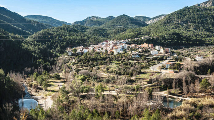 A hillside village nestled in a forested valley with a winding river in the foreground under a clear blue sky, mountains in the distance.