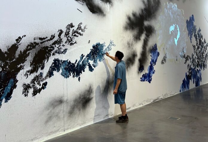 Man in a blue shirt and shorts paints a large abstract blue-black mural on a white wall.