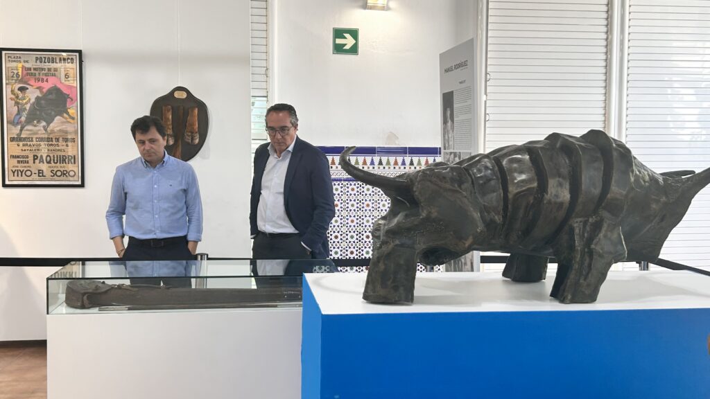 Two men in business attire stand beside a large bull sculpture in a museum gallery.