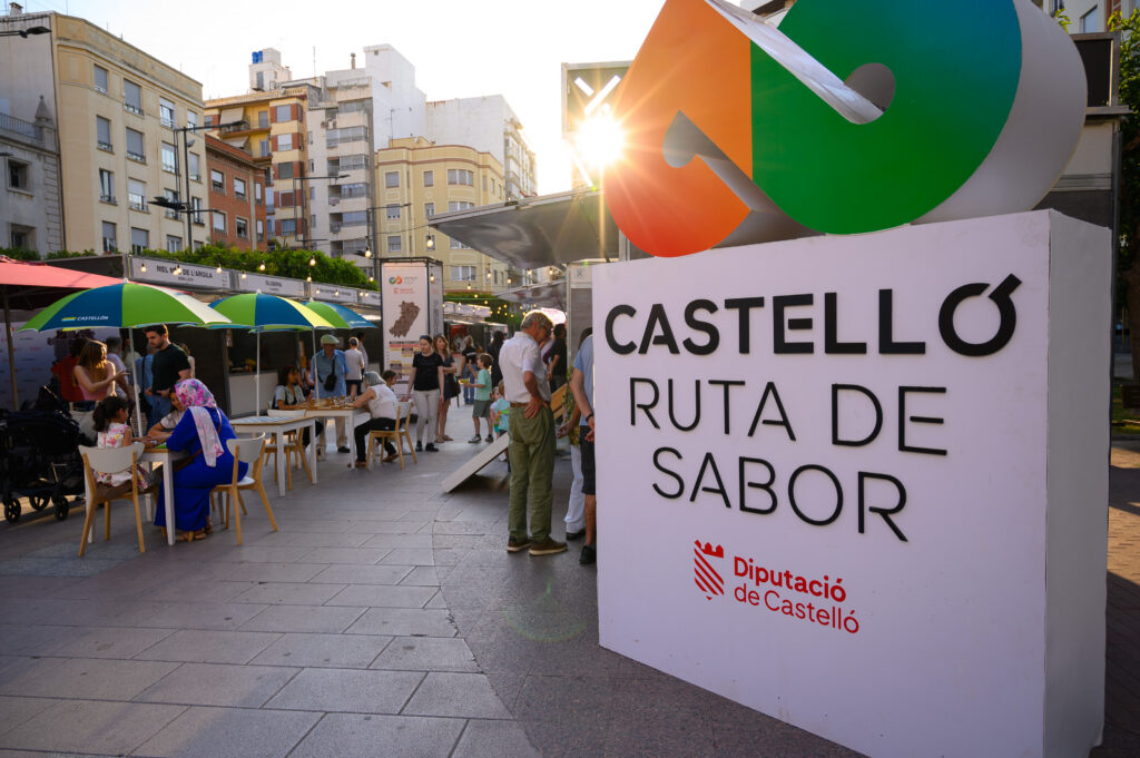 Outdoor market scene in Castelló with colorful umbrellas, people seated at tables, and a large sign reading CASTELLÓ RUTA DE SABOR.