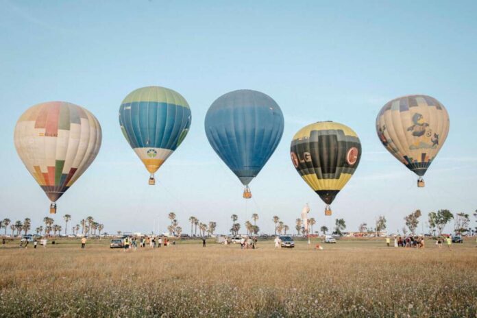 Five colorful hot air balloons lift off over a grassy field with spectators and cars nearby. Palm trees line the distant horizon at a clear day scene.