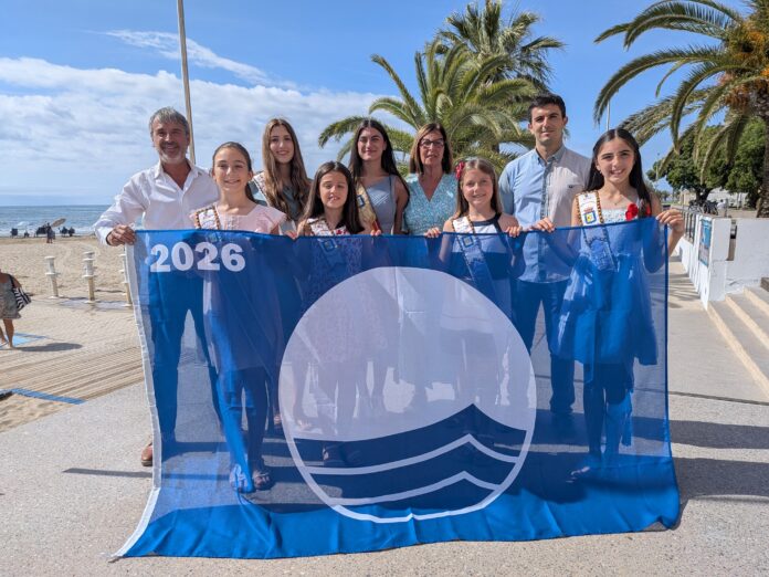 Group of eight people posing on a sunny beach promenade, holding a blue banner with a white circular emblem; ocean and palm trees in the background.