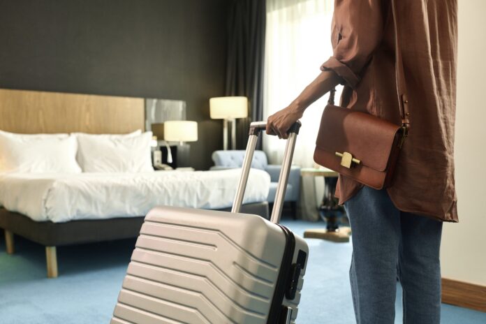 Traveler with a brown leather bag stands beside a rolling suitcase in a hotel room.