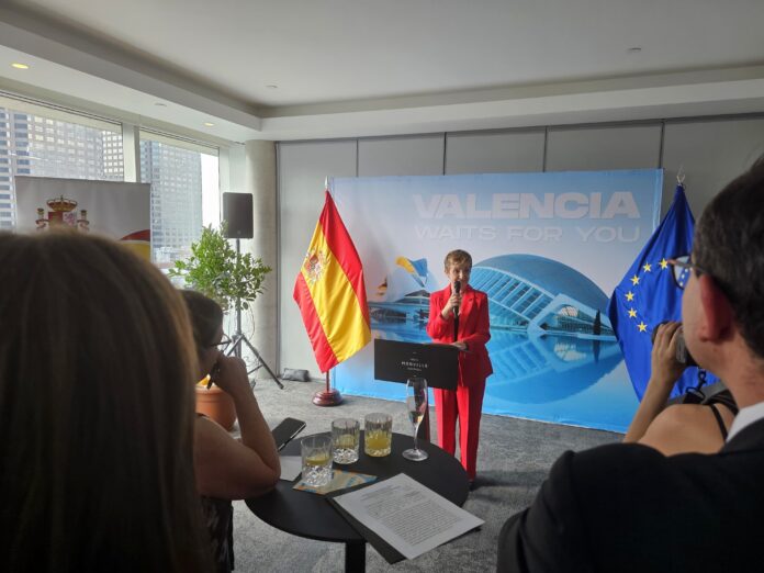 Speaker in a red suit presenting at a podium with a Valencia backdrop and Spanish flag beside him.