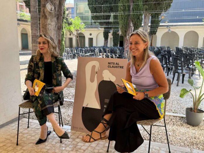 Two women sit on stools outdoors, holding yellow booklets, with a poster between them and many empty chairs in the background.