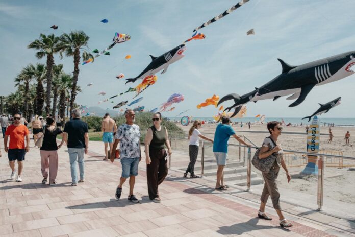 People stroll along a palm-lined beachfront boardwalk while giant shark-shaped kites soar overhead in the sky.