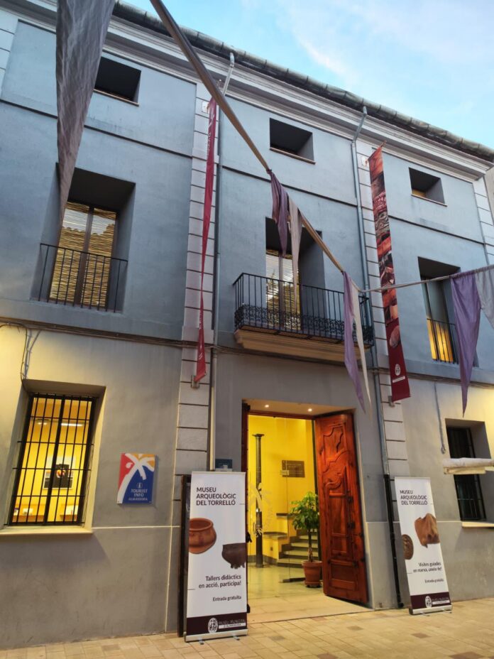 Facade of a pale blue building housing a museum, with colorful vertical banners and an open wooden door at the entrance
