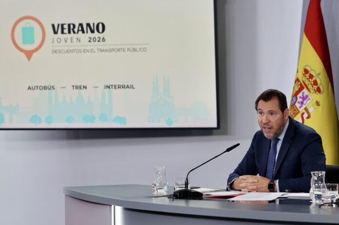 Man in a suit speaks at a conference table, a screen behind him shows 'VERANO JOVEN 2026' with a cityscape graphic and a Spanish flag nearby.