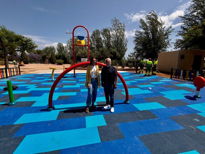 Two adults pose together on a bright blue playground surface with a red arched frame nearby, under a sunny sky.