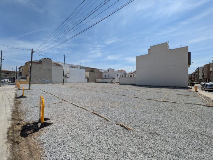 Empty gravel lot with yellow bollards and low-rise buildings along the edge, under a blue sky with power lines overhead
