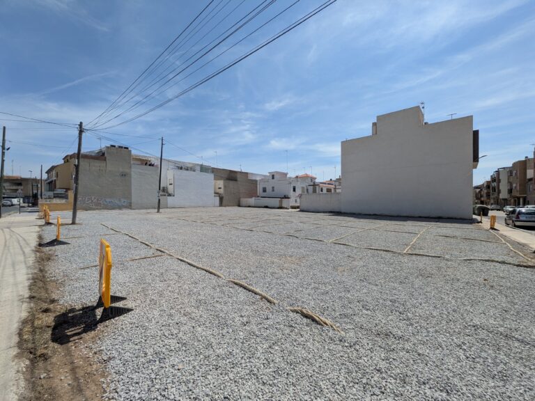 Empty gravel lot with yellow bollards and low-rise buildings along the edge, under a blue sky with power lines overhead