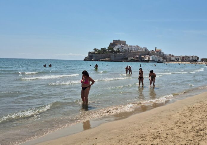 People wading in shallow water at a sunny beach with a hilltop castle town in the distance.
