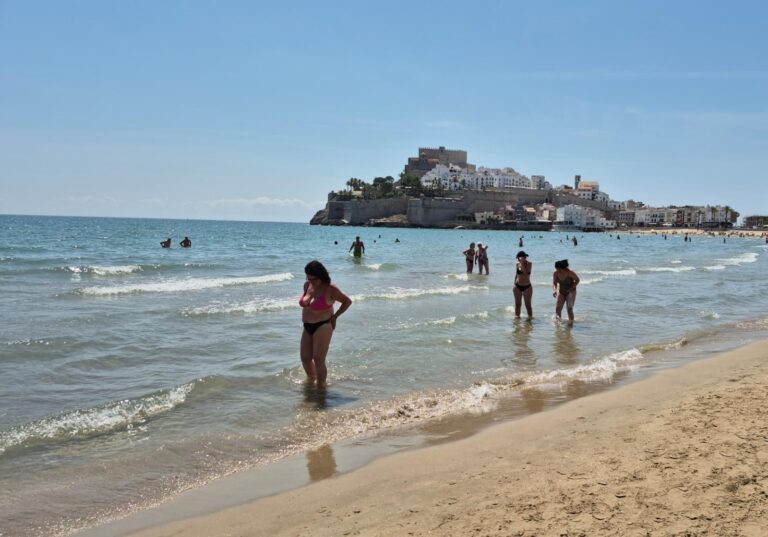 People wading in shallow water at a sunny beach with a hilltop castle town in the distance.