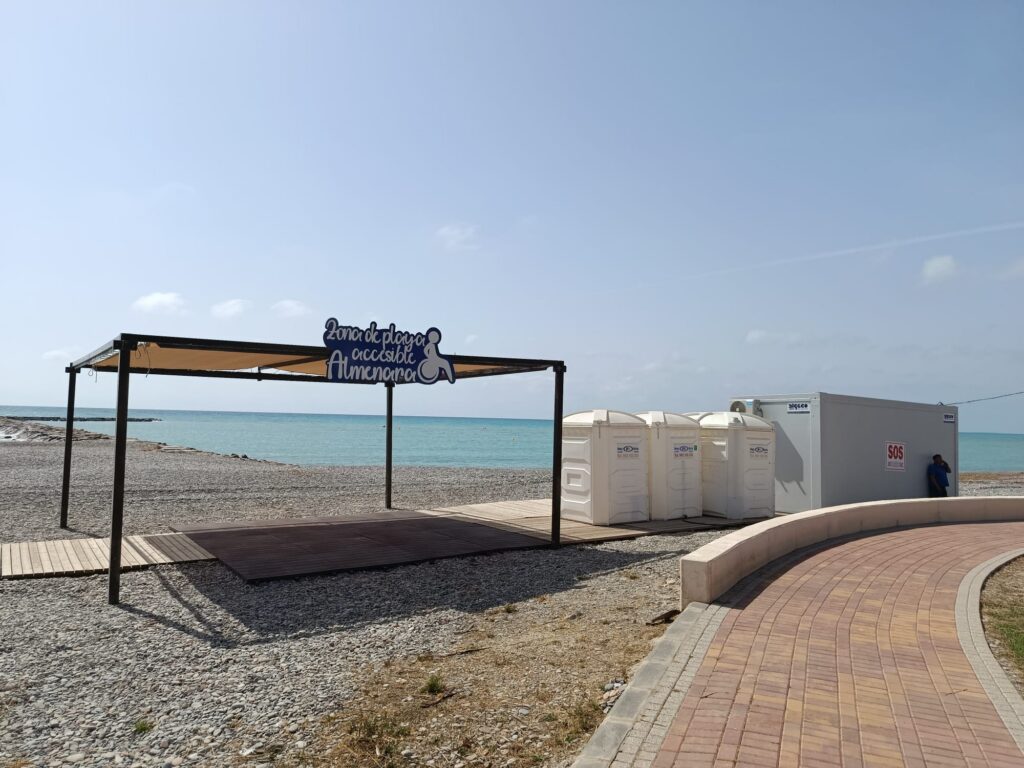 Beachfront scene with a metal frame canopy and a sign reading 'Zona de playa accesible' near the sea; portable toilets visible to the right under a clear blue sky.