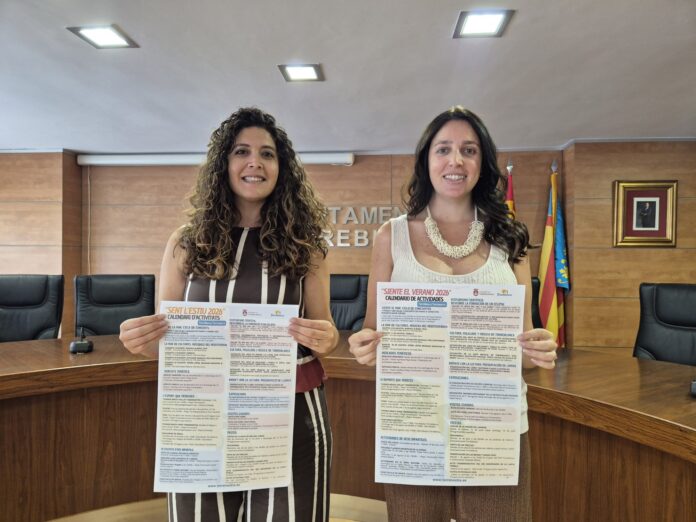 Two smiling women stand behind a wooden conference table, each holding large event flyers with dense text in Spanish.