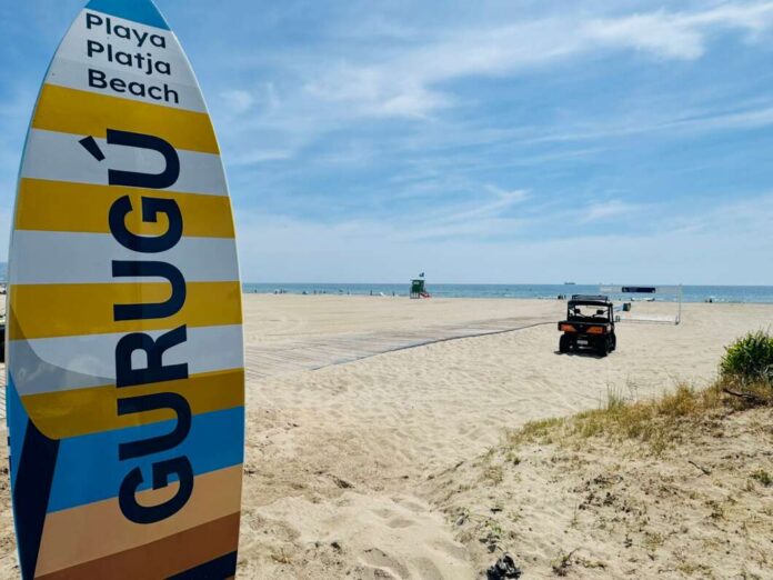 Colorful striped surfboard-shaped sign on a sandy beach with the sea in the distance and a small vehicle nearby left of center.