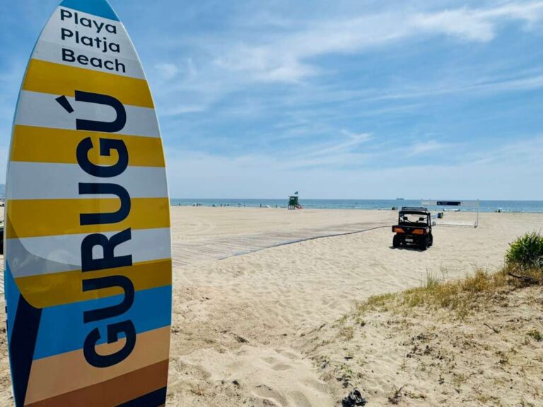 Colorful striped surfboard-shaped sign on a sandy beach with the sea in the distance and a small vehicle nearby left of center.