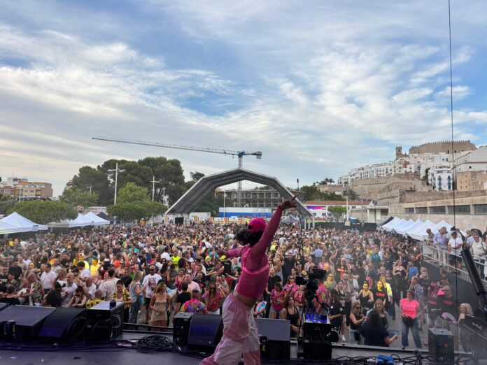 Large outdoor concert crowd facing a stage with a performer in a bright pink outfit raising an arm.