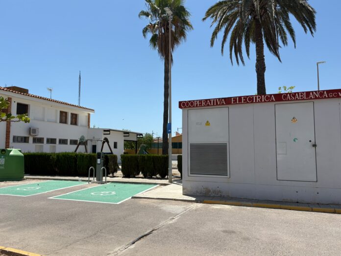 Exterior view of a small electrical utility building with a red sign reading 'Cooperativa Electrica Casablanca' beside tall palm trees against a blue sky; green-marked accessible parking spots in foreground.