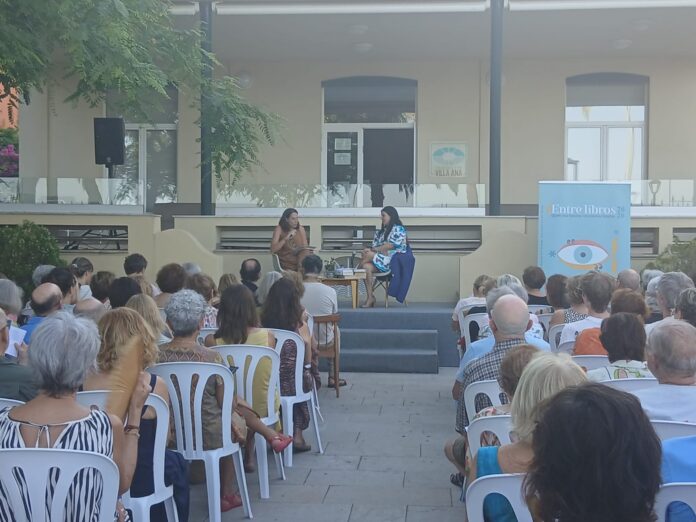Two women sit on a small stage and talk, an outdoor book event with a seated audience nearby.
