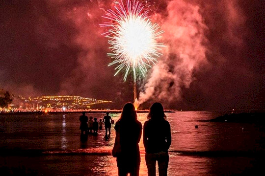 Two silhouetted people stand at the water's edge watching a bright fireworks display over a dark lake at night.