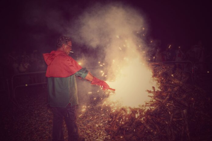 Man in a red and teal jacket and gloves tending a large bonfire at night, sparks flying around.