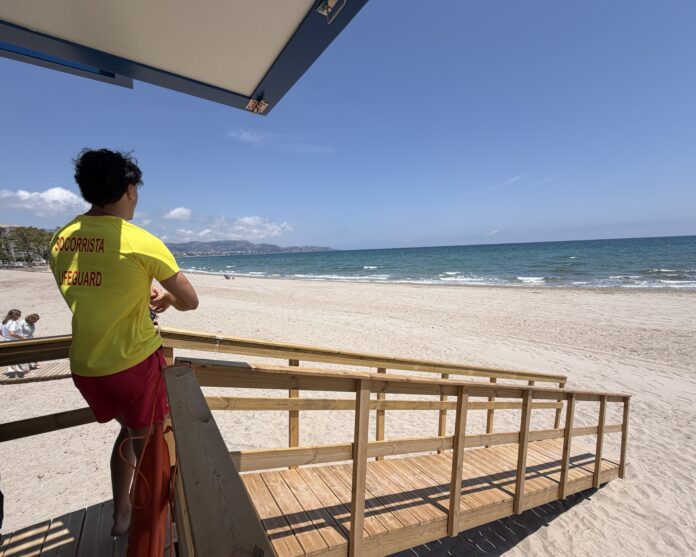 Lifeguard in a yellow shirt standing on a wooden viewing deck at the sunny beach, facing the sea.
