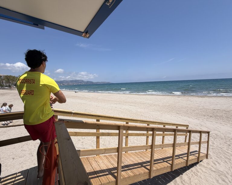 Lifeguard in a yellow shirt standing on a wooden viewing deck at the sunny beach, facing the sea.