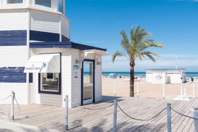 White beachside information booth with blue trim and a 'Tourist Info' awning on a wooden boardwalk, palm tree, and sandy beach in the background.