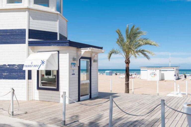 White beachside information booth with blue trim and a 'Tourist Info' awning on a wooden boardwalk, palm tree, and sandy beach in the background.