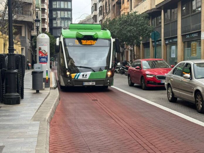 Green tram in a city street moving on a dedicated red-brick track with cars nearby on the right side of the road.