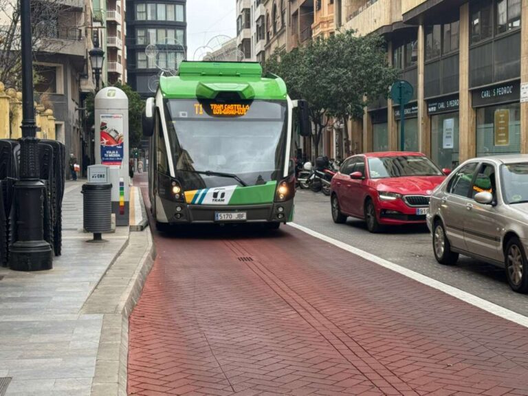 Green tram in a city street moving on a dedicated red-brick track with cars nearby on the right side of the road.