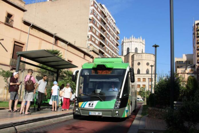 Front view of a green tram arriving at a stop, with several people waiting on the platform under a shelter, in an urban street with buildings and a church in the background.