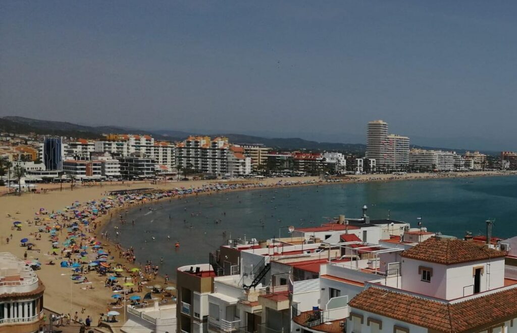 Busy sandy beach with colorful umbrellas and sunbathers along a curved coastline, with modern buildings in the background.