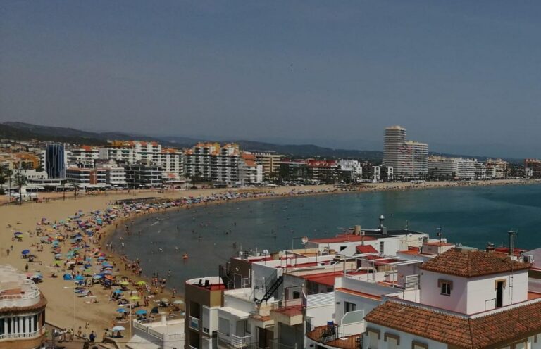Busy sandy beach with colorful umbrellas and sunbathers along a curved coastline, with modern buildings in the background.