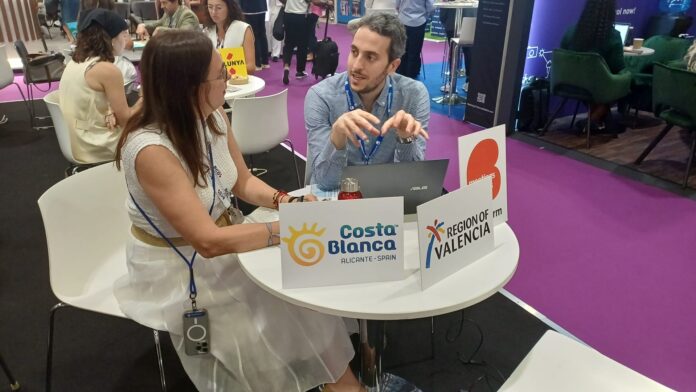Two professionals at a trade-show booth, talking across a round white table with Costa Blanca Alicante Spain and Region of Valencia signs.