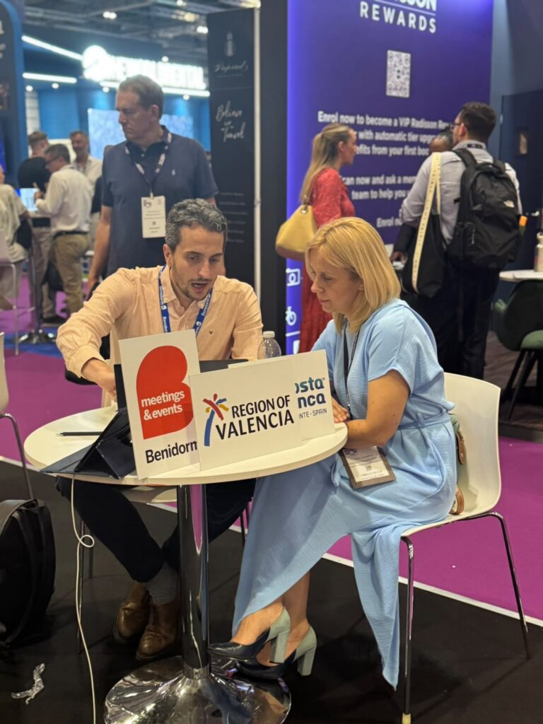 Two professionals sit at a round table in a conference hall, examining travel signs for Valencia and Benidorm with 'Region of Valencia' branding displayed on the signs.