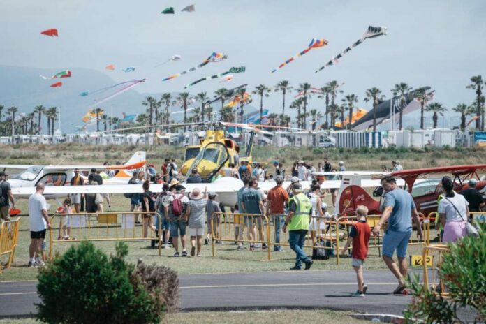 Crowd gathered behind barriers at an air show, with a yellow helicopter and small planes on display and colorful kites in the sky.