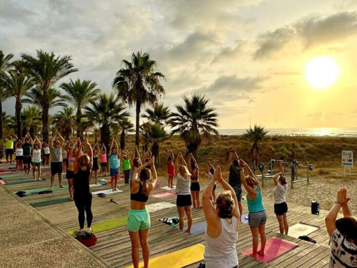 Group of people practicing yoga on colorful mats on a wooden deck by the beach at sunset, with palm trees in the background.
