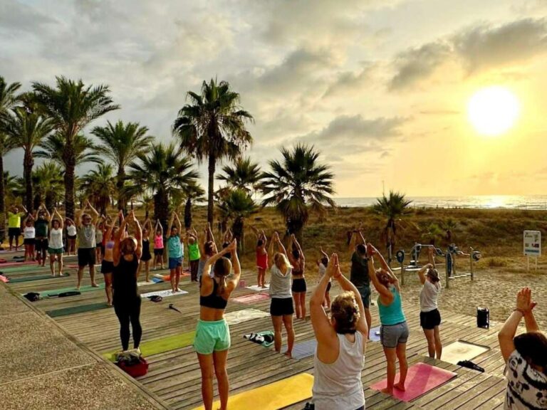 Group of people practicing yoga on colorful mats on a wooden deck by the beach at sunset, with palm trees in the background.