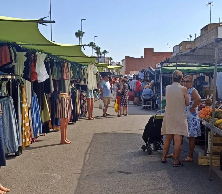 Outdoor market street with clothing stalls on both sides and shoppers browsing under green canopies.