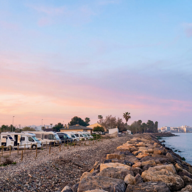 Rocky beach at sunset with a line of camper vans parked along a path and a wooden railing, palm trees in the distance, and calm water on the right.