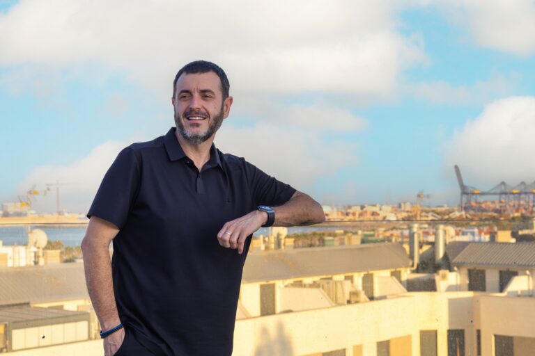 Smiling man in a black polo leaning on a glass railing with a city skyline and blue sky behind him.