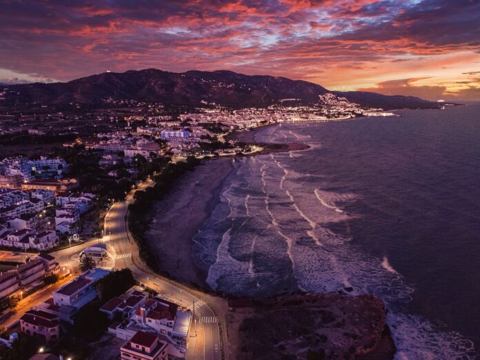 Aerial view of a coastal town at sunset, with a purple-orange sky, streetlights along the curved shoreline and waves.