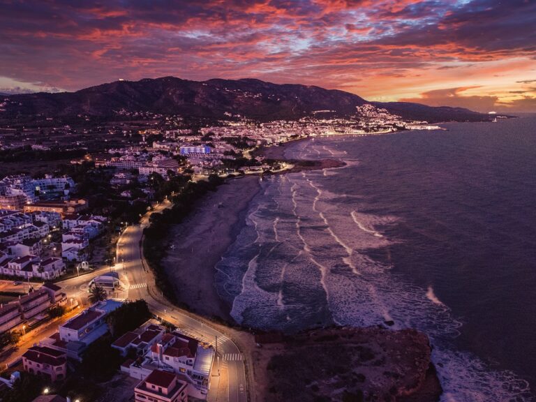 Aerial view of a coastal town at sunset, with a purple-orange sky, streetlights along the curved shoreline and waves.