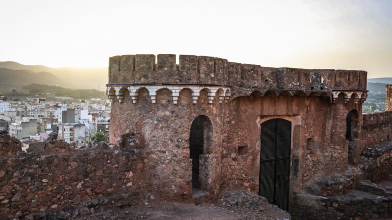 Ancient stone fortress wall with crenellations overlooking a city at sunset.