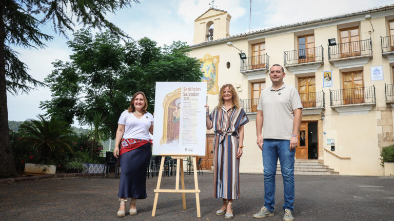Three adults stand beside an easel with a tall poster about Santíssim Salvador in front of a pale building and greenery behind them on a sunny day.