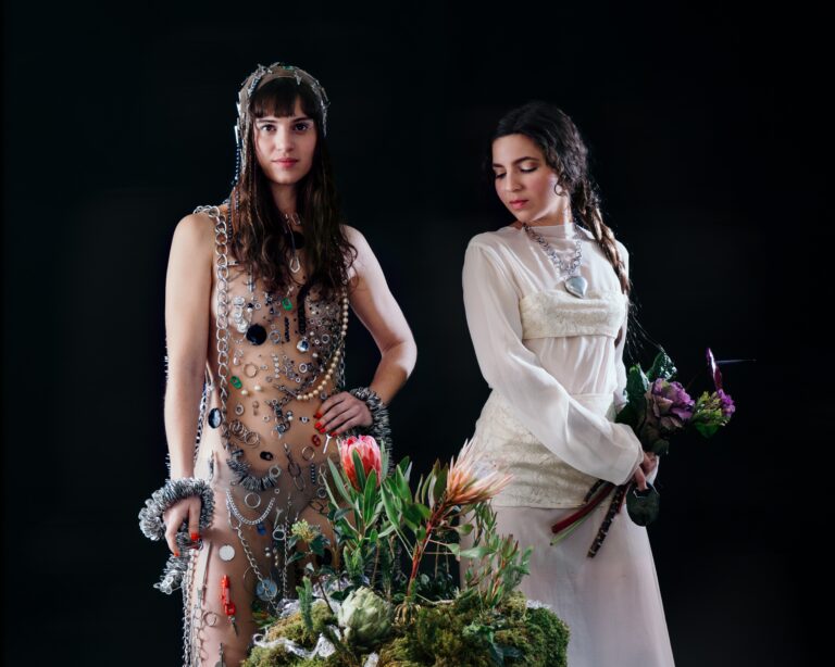 Two women pose against a dark background: one covered in metallic chains and jewelry, the other in a cream dress holding flowers.