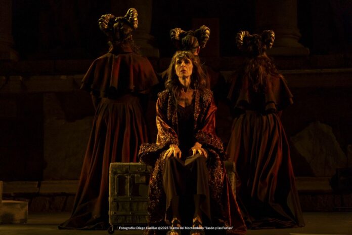 Seated woman in a sparkling burgundy robe on a dark stage, flanked by two robed figures with tall, horned headpieces.