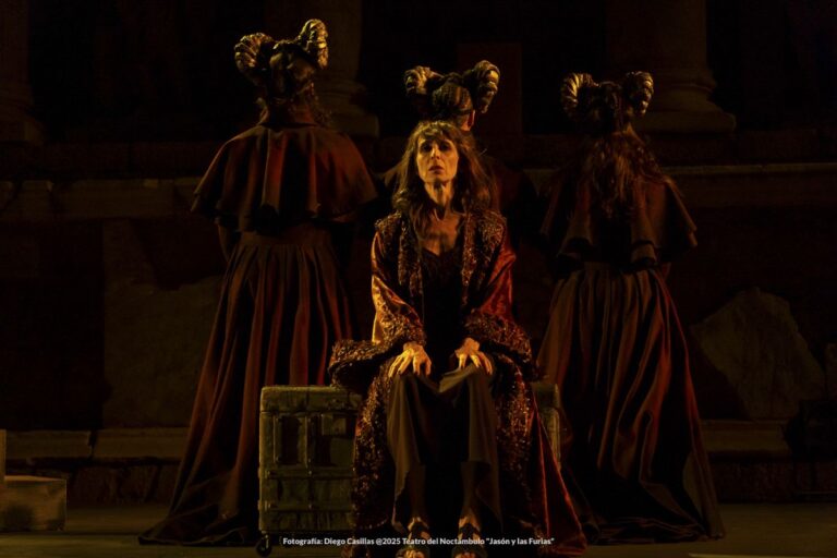 Seated woman in a sparkling burgundy robe on a dark stage, flanked by two robed figures with tall, horned headpieces.
